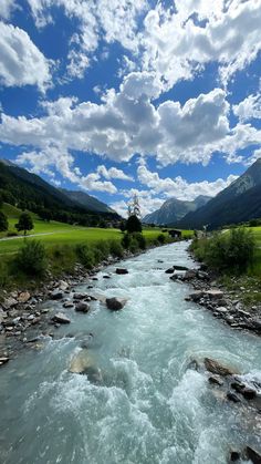 a river running through a lush green valley under a blue sky with white fluffy clouds