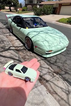 a hand holding a toy car in front of a small model car on the street