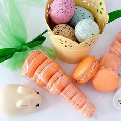 an assortment of colorful macaroons in a basket next to some bunny ears and carrots