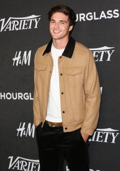 a man standing in front of a black background wearing a tan jacket and white shirt