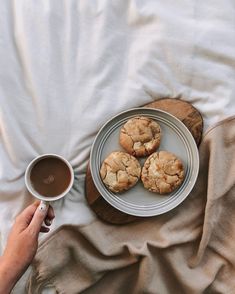 a person holding a cup of coffee next to some cookies on a plate and blanket