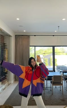 a woman standing in a living room holding her arms out to the side while wearing a colorful jacket and white pants