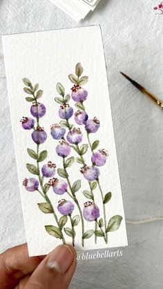 a hand holding up a card with watercolor flowers on it, next to some paintbrushes