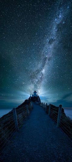 the night sky is filled with stars above a wooden walkway leading to a light house