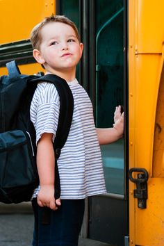 a little boy standing in front of a school bus with his back pack on the door