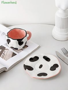 a white table topped with a black and white cow shaped coffee cup next to a plate