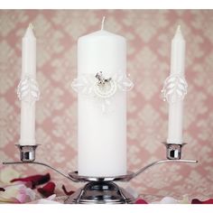 three white candles are sitting on a silver candle holder with flowers around it and pink wallpaper in the background