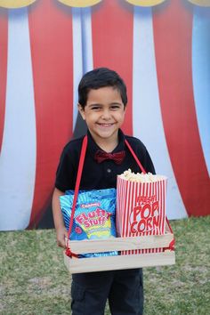 a young boy holding a box of popcorn and two bags of chips in front of a circus tent