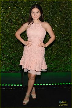 a young woman in a pink dress poses for the camera on a black carpet with bushes behind her