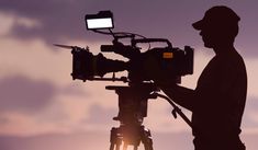 a man standing next to a camera on top of a tripod in front of a cloudy sky