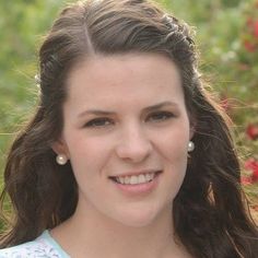 a woman with long brown hair wearing a blue top and pearl earrings smiling at the camera