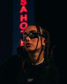 a woman with dreadlocks wearing sunglasses in front of a neon sign