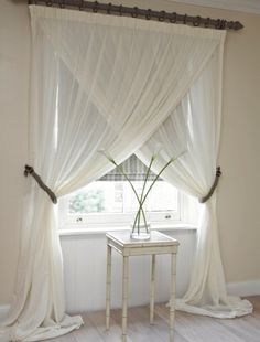 a white table sitting under a window next to a sheer curtain with flowers in it