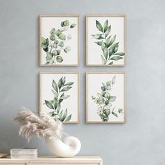 three green leaves are hanging on the wall next to a white vase and two framed pictures