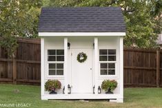 a small white shed with potted plants and wreath on the door is shown in front of a fence