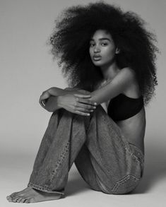 a woman with an afro sitting on the ground wearing jeans and a tank top, looking off to the side