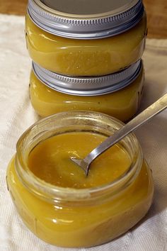 three jars filled with yellow liquid sitting on top of a white cloth next to a spoon