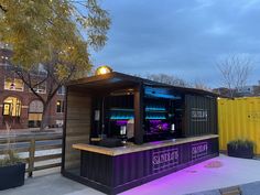 an outdoor bar is lit up with purple lights and sits next to a yellow fence