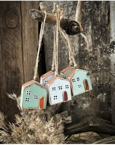 three small houses hanging from strings on a wooden wall with dried grass in the foreground