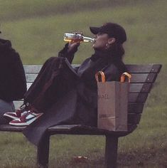 two people sitting on a park bench drinking beer and eating food from paper bags in front of them