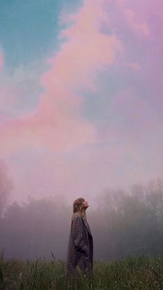 a woman standing in the middle of a field looking up at a pink cloud filled sky