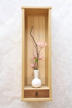 a white vase with pink flowers in it sitting inside a wooden box on the wall