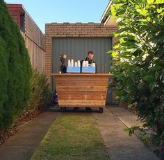 a man standing behind a wooden counter in front of a building with trees and bushes