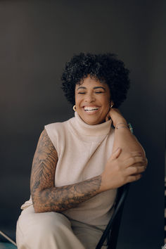 a woman sitting in a chair smiling and looking off to the side with tattoos on her arm