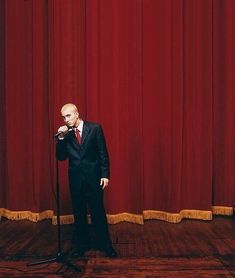 a man standing in front of a microphone on top of a hard wood floor next to a red curtain