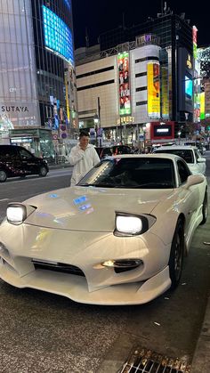 a man standing next to a white sports car in the middle of a city at night