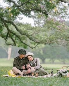 a man and woman sitting on the ground under a tree with wine glasses in their hands