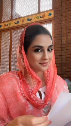 a woman wearing a pink shawl sitting in front of a window and holding a piece of paper