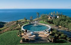 an outdoor swimming pool surrounded by lawn chairs and trees with the ocean in the background