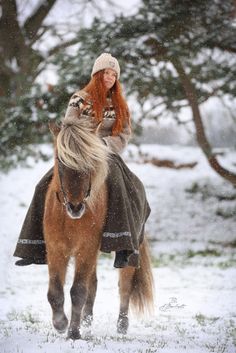 a woman riding on the back of a brown horse through snow covered ground next to trees