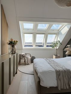 a bed sitting under a window in a bedroom next to a chair and table with vases on it