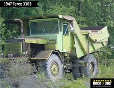 an old green dump truck is parked in the grass near some trees and bushes,