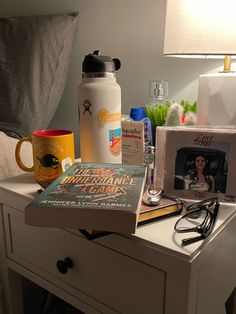 a table topped with books and other items on top of a white dresser next to a lamp