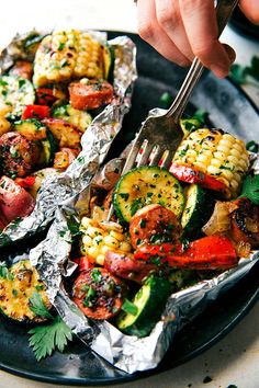 a person is holding a fork over some foil with vegetables on it and other food in the background