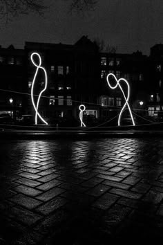 two figures are lit up in the dark on a brick street at night with buildings behind them