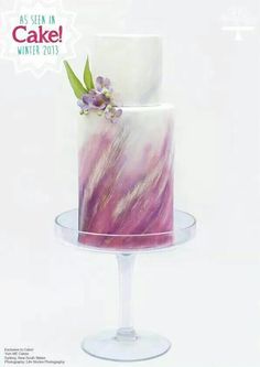 a three tiered cake with purple and white frosting on a clear pedestal, decorated with flowers