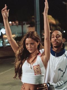 a man and woman standing next to each other on the street at night with their arms in the air