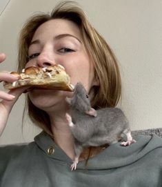 a woman biting into a piece of pizza with a rat on her shoulder next to her