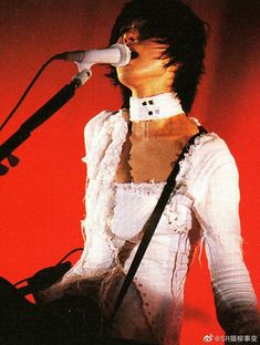 a woman singing into a microphone while standing in front of a red wall and holding a guitar