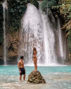 two people are standing in the water near a waterfall and one person is looking at it