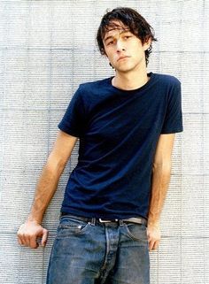 a young man leaning against a wall with his hands in his pockets and looking at the camera