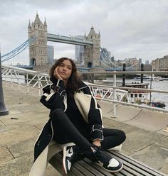 a woman sitting on top of a wooden bench next to a river and bridge in the background