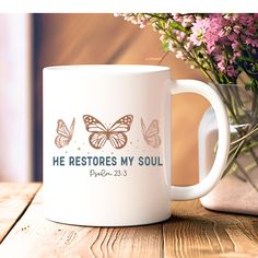 a white coffee mug with the words he restores my soul printed on it next to a bouquet of flowers