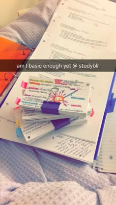 a pile of papers sitting on top of a bed next to an orange book and pen