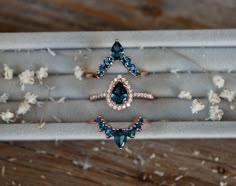 three rings with blue and white stones in a box on top of wooden table next to dried flowers