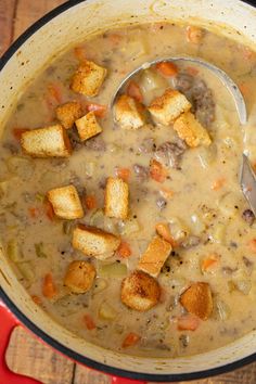 a red pot filled with soup and croutons on top of a wooden table